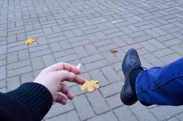 Un hombre sostiene un cigarrillo en sus manos, dedos sentados en el parque y fumando con su pierna en un zapato sobre el fondo de losas de pavimentación con hojas de otoño. El trasfondo — Foto de Stock