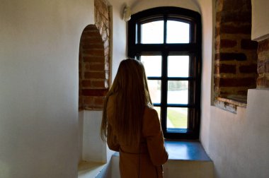 Bir kız, bir kadın bir eski antik taş beyaz kalın beton tuğla duvar eski kalede bir çerçeve ile a küçük dar siyah pencere önünde duruyor. Arka plan