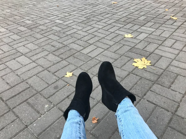 Pernas finas femininas em jeans e botas, sapatos contra um fundo de lajes de pavimentação de concreto de pedra cinza e folhas amarelas de outono. O fundo — Fotografia de Stock