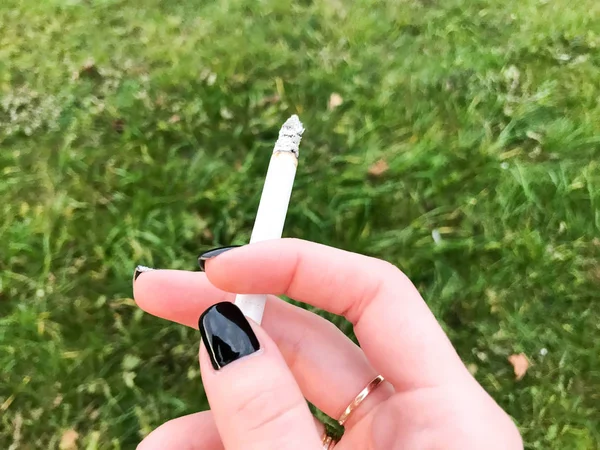The hand of a smoking girl, a woman with beautiful fingers and a gold wedding ring and black manicure on the nails holding a burning cigarette in the background of green grass — Stock Photo, Image