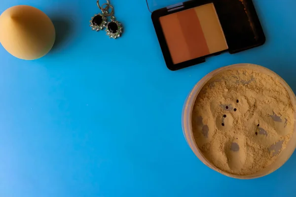 Beauty box, loose matte mineral powder and blush with a beauty blender for makeup and silver earrings on a blue background. Flat lay. Top view