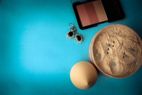 Beauty box, loose matte mineral powder and blush with a beauty blender for makeup and silver earrings on a blue background. Flat lay. Top view