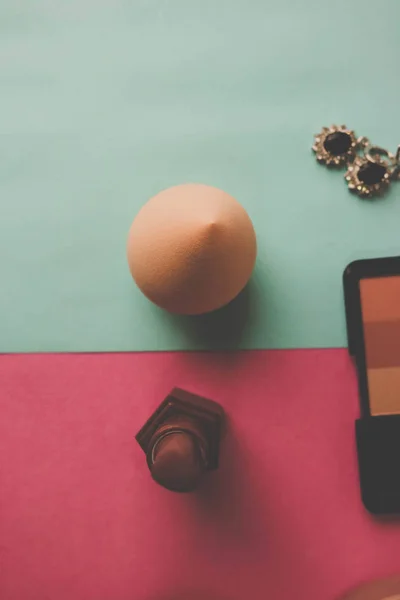 Beauty box, makeup kit, powder and blush with beauty blender, perfume, lipstick, highlighter and earrings for makeup on a pink purple and blue background. Flat lay. Top view