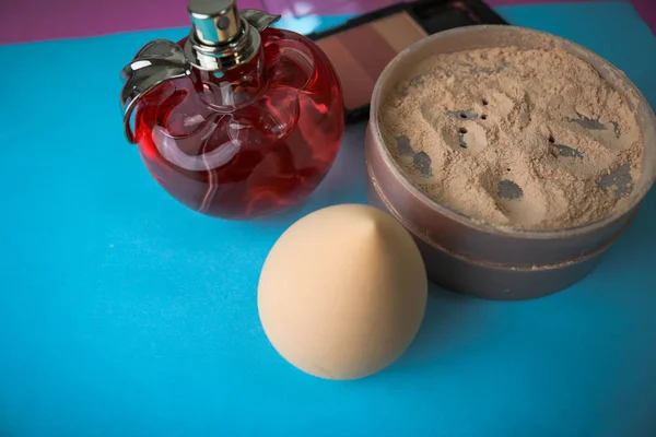 Beauty box, loose matte mineral powder and blush with a beauty blender for makeup and red glass bottle of perfume on a blue background. Flat lay. Top view