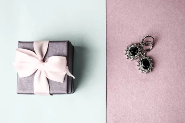 Beauty box, festive beautiful gift box with a bow with silver earrings with precious stones on a pink purple and blue background. Flat lay. Top view