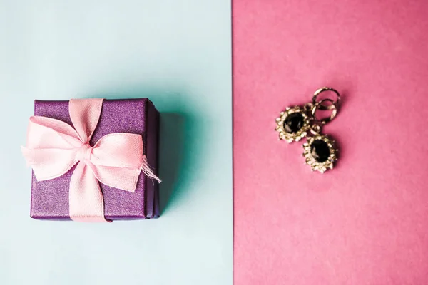 Beauty box, festive beautiful gift box with a bow with silver earrings with precious stones on a pink purple and blue background. Flat lay. Top view