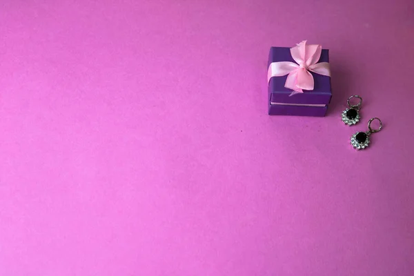 Beauty box, festive beautiful gift box with a bow with silver earrings with precious stones on a pink purple background. Flat lay. Top view