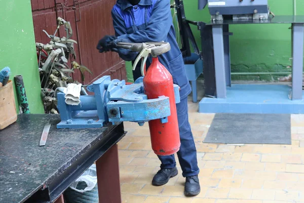 Um homem trabalhando em um grande vício industrial de metal com uma chave de gás enorme desaperta uma porca em um cilindro extintor vermelho em uma oficina de fábrica — Fotografia de Stock