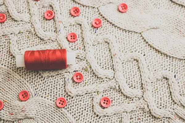 Beautiful texture of a soft warm natural sweater, fabrics with a knitted pattern of yarn and red small round buttons for sewing and a skein of red thread. The background — Stock Photo, Image