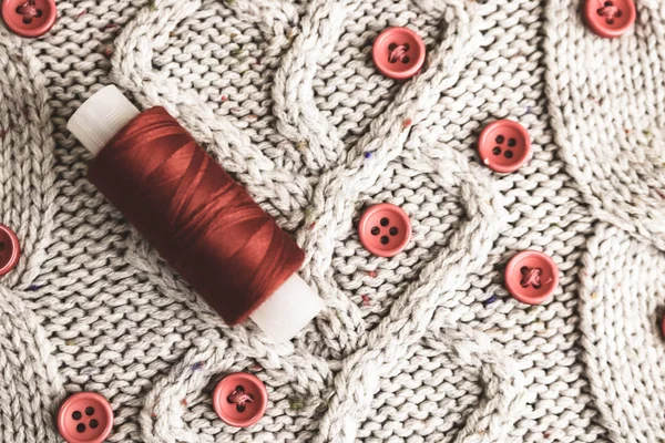 Beautiful texture of a soft warm natural sweater, fabrics with a knitted pattern of yarn and red small round buttons for sewing and a skein of red thread. The background — Stock Photo, Image