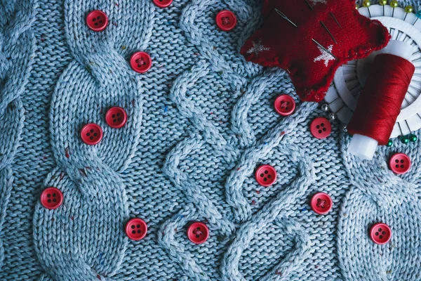 Beautiful texture of a soft warm natural sweater, fabrics with a knitted pattern and red small round buttons for sewing and a skein of thread, needle bed and needle pad. Flat lay. The background — Stock Photo, Image