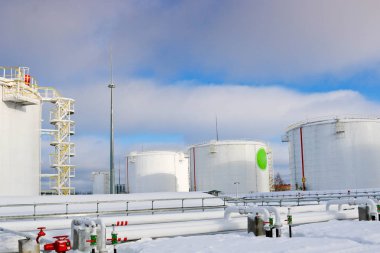 Large white iron metal industrial tanks for storage of fuel, gasoline and diesel and pipeline with valves and flanges at the refinery in the winter