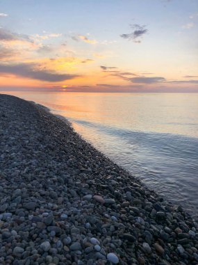 Deniz, nehir, göl, gölet, turuncu gökyüzüne karşı yuvarlak taşlar ve su ile tropikal sıcak bir tatil kayalık plajda güzel sarı parlak gün batımı. Dikey görünüm