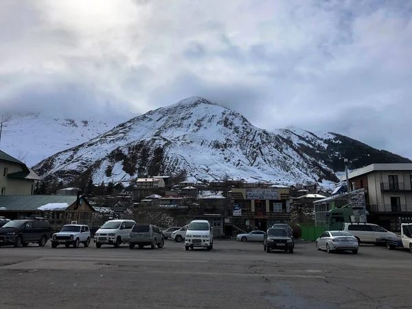 駐車場や石造りの家、建物、ショップ、青い空に対して雪で覆われた高い山頂と岩と美しい山の冬のリゾートに給油。ジョージア、トビリシ、2019年4月17日 — ストック写真