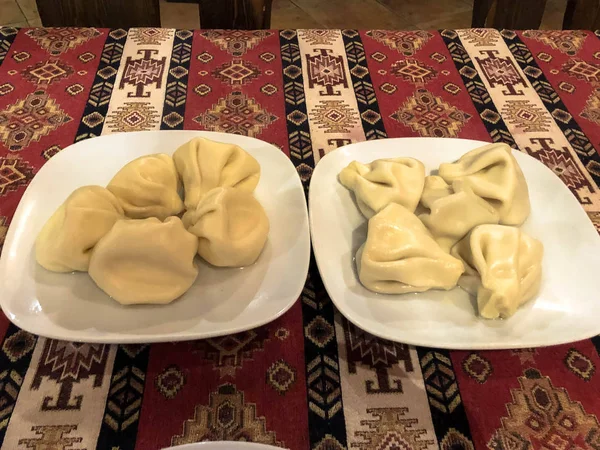 Duas porções de khinkali saboroso nutritivo, manti em placas brancas de cerâmica na mesa no restaurante georgiano nacional na mesa com uma toalha de mesa vermelha — Fotografia de Stock