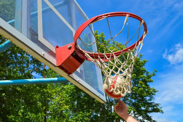 Esportes vermelhos cesta redonda basquete streetball na área de rua aberta sob o céu aberto com uma rede contra o céu azul e árvores e a mão de um atleta com a bola — Fotografia de Stock