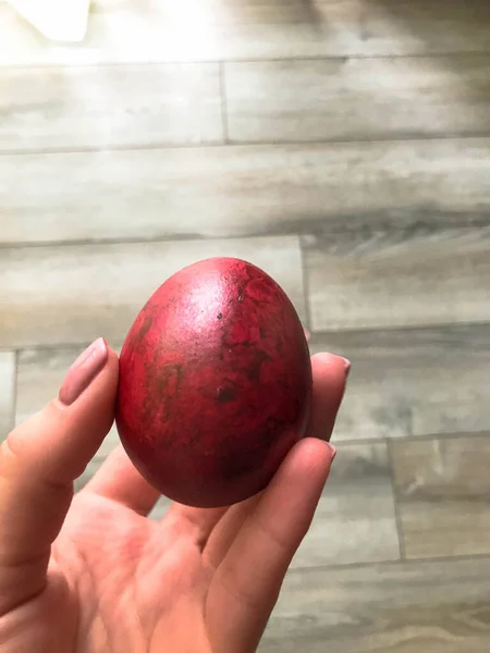 Easter egg of red color in female hands with pink cute manicure. paint the shell with onion peel. brilliant decoration of the Easter basket. on a wood background — Stock Photo, Image