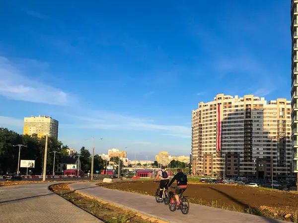 Ciclistas viajan a lo largo del carril bici en el asfalto con el telón de fondo de un edificio residencial. una bandera blanca-roja-blanca cuelga en la casa en una forma inusual diadema —  Fotos de Stock