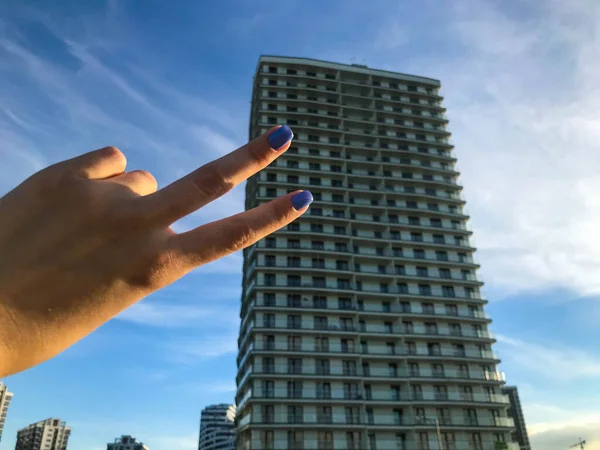 Edifício alto apartamento branco, edifício de concreto. arranha-céu com janelas panorâmicas, em estilo árabe com terraços. menina segurando os dedos em um sinal de paz. manicure criativo azul em pregos — Fotografia de Stock