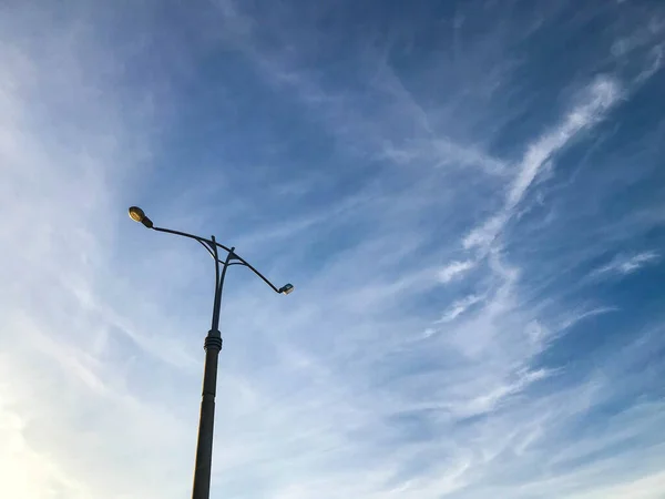 Laternenpfahl mit weißer Glühbirne vor klarem Himmel. Laterne, um zwei Straßen zu beleuchten. graue Säule mit Laterne, warmes Gelb. neben der Spur eines fliegenden Flugzeugs — Stockfoto