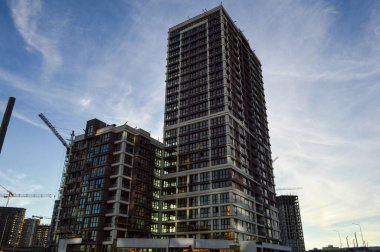 Şehirde yüksek ve çok katlı binalar yan yana durur. Beyaz-kahverengi renkli cam cepheler. Evlerin panoramik pencereleri var. Yakınlarda bir vinç var.