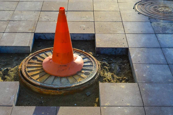 a bright orange cone stands on the sewer drain. marking of dangerous sections on the road, entry for people is prohibited. the cone stands on the broken asphalt, the builders are laying a new surface