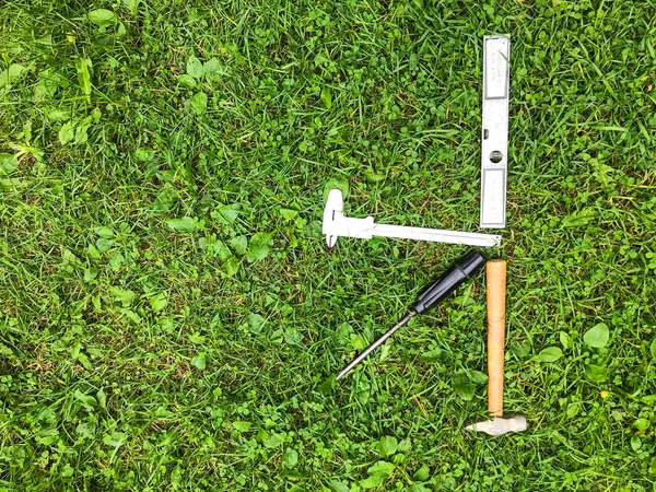 Na trawie leży młotek i śrubokręt, poziom i inne. Narzędzia naprawcze w kształcie litery K znajdują się na trawniku. Młot na drewnianej rączce, śrubokręt z czarną rączką — Zdjęcie stockowe