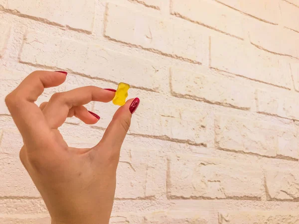 A gummy yellow bear in the hands of a girl with a red manicure. bear are kept on a white brick background. homemade wall with sweet candy. mouth-watering dessert — Stock Photo, Image