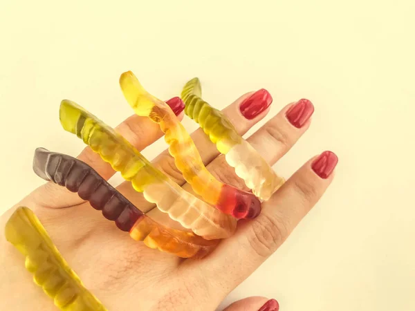 A lot of gummy worms lie with a girl with a red manicure on an orange matte background. delicious appetizing dessert. bright gummy worms, sweet gelatinous candies — Stock Photo, Image