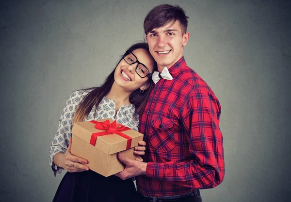Young content couple embracing and standing with gift box having anniversary celebration and looking at camera