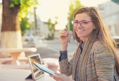 Oldukça şık hipster kadın tablet ve çalışmaları olan ve kameraya gülümseyen Not defteri ile sokakta oturan gözlüklü