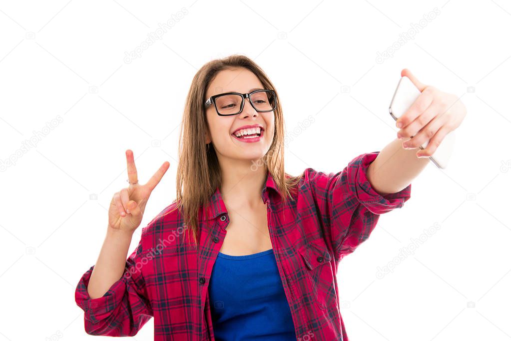 Charming young modern woman showing v-sign and taking selfie with smartphone isolated on white background