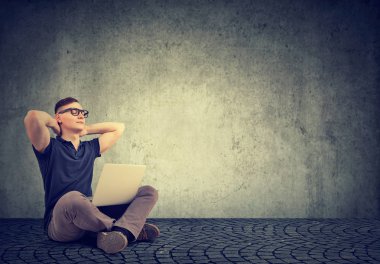 Genç casual adam ile laptop gri arka plan üzerinde oturan ve rahatlatıcı elele başının arkasında gözlük