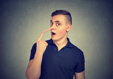 Young man looking surprised at camera touching long fake nose of liar on gray background clipart
