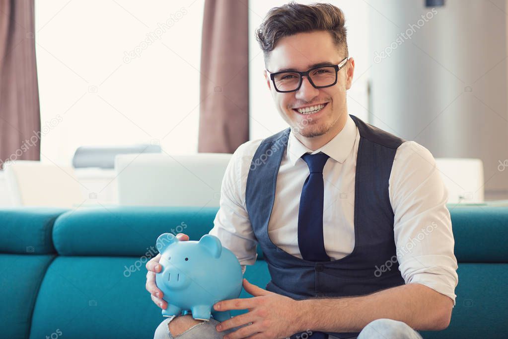 Happy formal man in glasses holding blue piggy bank sitting on sofa in new modern apartment smiling at camera