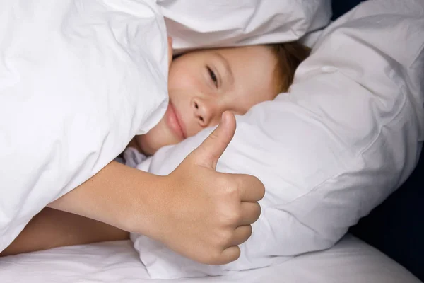 Senyum anak laki-laki terletak di tempat tidur dengan linen tempat tidur putih dan menunjukkan jempol up — Stok Foto