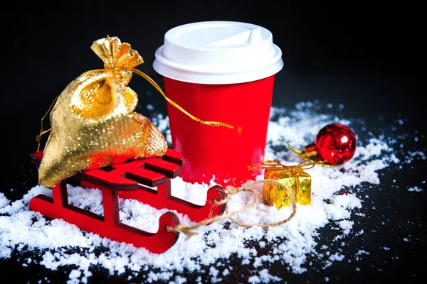 Café para ir em copo vermelho, trenó, saco de ouro e natal presente no fundo preto. Bebidas quentes no inverno frio . — Fotografia de Stock