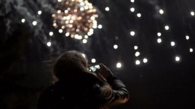 Kız bir cep telefonu üzerinde havai fişek çekiyor. Arka planda gökyüzünün ışıklar tarafından aydınlatılmış siluet.