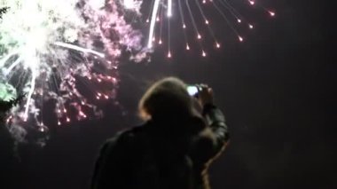 Kız bir cep telefonu üzerinde havai fişek çekiyor. Arka planda gökyüzünün ışıklar tarafından aydınlatılmış siluet.
