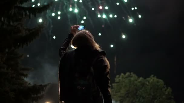 La chica toma fotos de fuegos artificiales en un teléfono móvil. Silueta en el fondo del cielo iluminada por luces . — Vídeos de Stock