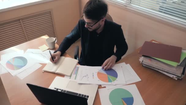 Fotografía media de un joven exitoso trabajando en un ordenador portátil y escribiendo en un cuaderno — Vídeos de Stock