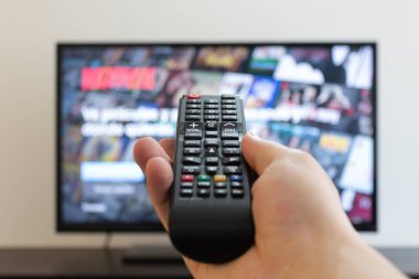 Arka planda bulanık bir televizyonu olan siyah bir televizyon kumandasını tutan bir erkeğe yakın çekim. İç mekan ve girişimcilik konsepti fotoğrafçılığı
