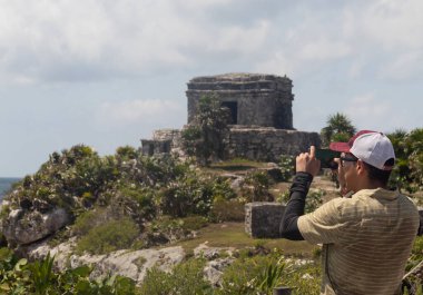Telefonuyla fotoğraf çeken genç bir adam Tulum arkeolojik bölgesine güneşli bir günde ünlü antik Maya rüzgar tanrısı tapınağına
