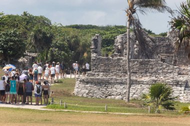 Bir grup ziyaretçi, güneşli bir günde antik bir Maya yapısının yakınından geçerek Tulum arkeolojik bölgesini harabeye çeviriyor.