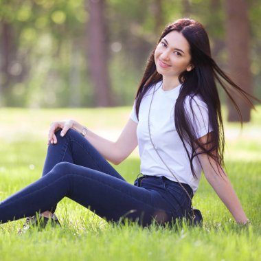 Genç kadın yeşil çimlerin üzerinde parkta dinleniyor. Renkli arka planlı güzel doğa sahnesi, yaz mevsiminde ağaçlar. Açık hava yaşam tarzı. Mutlu gülümseyen kadın yeşil çimlerde oturuyor.