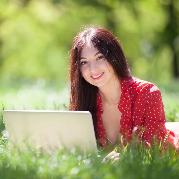 Mladá žena s bílým laptopem v parku. Krásná přírodní scéna s barevným pozadím, stromy v letní sezóně. Venkovní životní styl. Šťastná usměvavá žena s počítačem ležela na zelené trávě — Stock fotografie