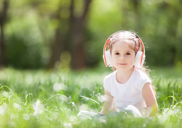 Söt liten flicka lyssnar på musik i parken. Familj utomhus livsstil. Glad liten unge i hörlurar sitter på grönt gräs. Skönhet på sommaren. Barndoms lycka. Barnens dag. — Stockfoto