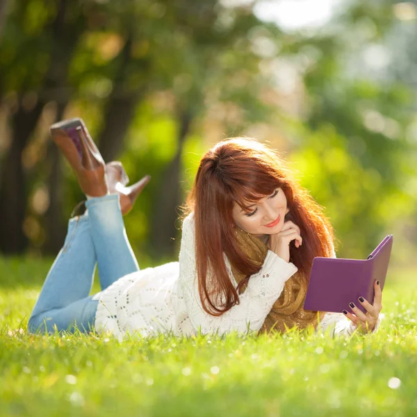 Joven mujer bonita leer libro electrónico en el parque. Belleza escena de la naturaleza con fondo de primavera colorido. Estilo de vida exterior. Feliz mujer sonriente relajarse con la tableta en la hierba verde — Foto de Stock