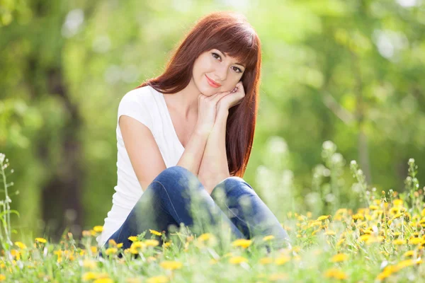 Młoda kobieta zrelaksować się w parku z kwiatami. Piękna scena przyrody z kolorowym tłem, drzewami i kwiatami w sezonie letnim. Styl życia na świeżym powietrzu. Szczęśliwy uśmiechnięta kobieta siedzi na zielonej trawie — Zdjęcie stockowe