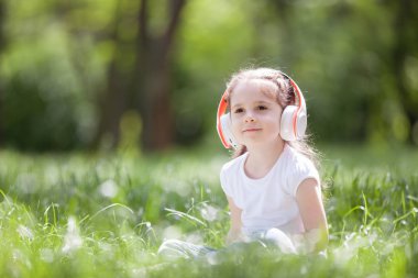 Şirin küçük kız parkta müzik dinliyor. Aile açık hava yaşam tarzı. Yeşil çimlerin üzerinde oturan kulaklıklı mutlu küçük çocuk. Yazın güzellik doğası. Çocukluk mutluluğu. Çocuk günü.